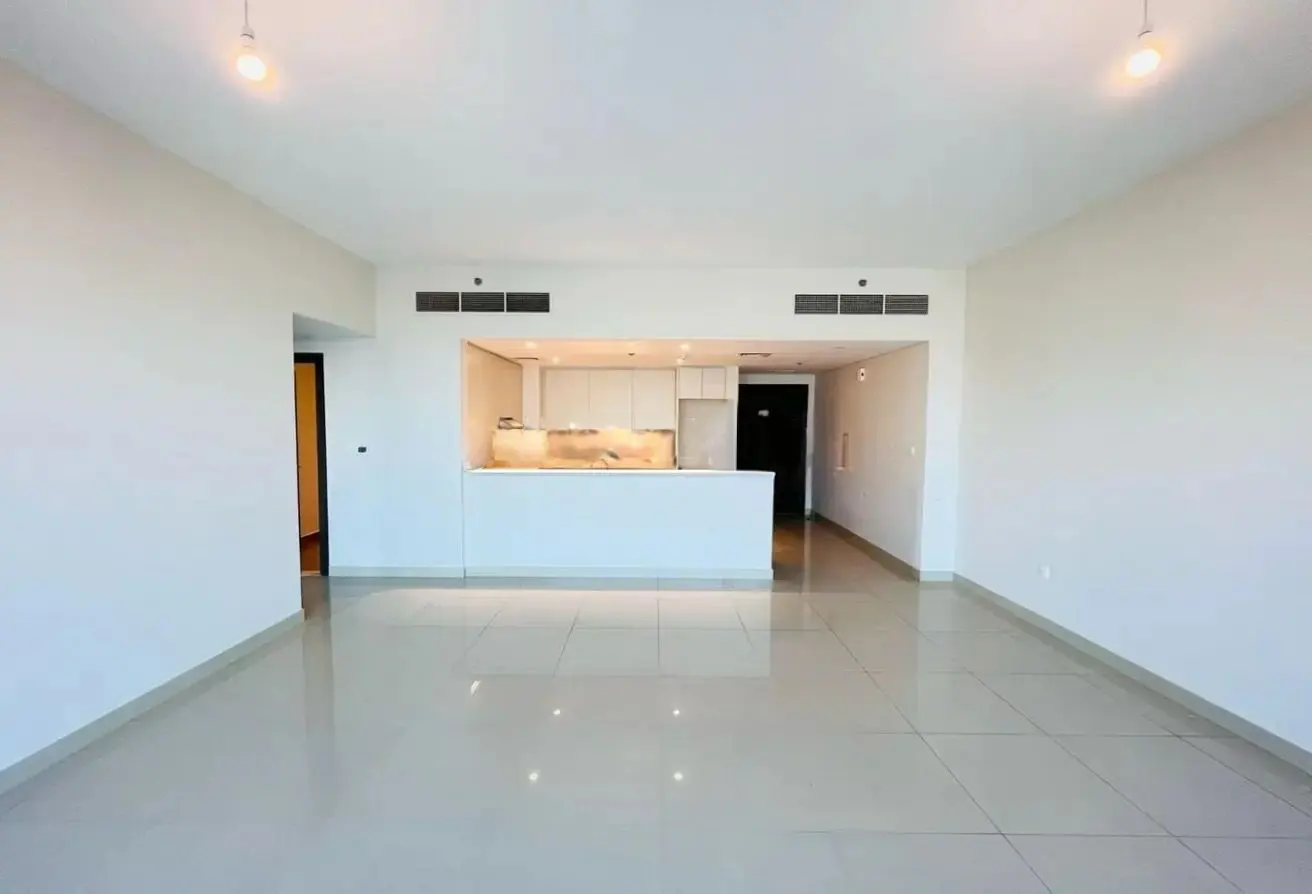 A bright, empty room with white walls and tile floors. An open kitchen with white cabinets is visible in the background.
