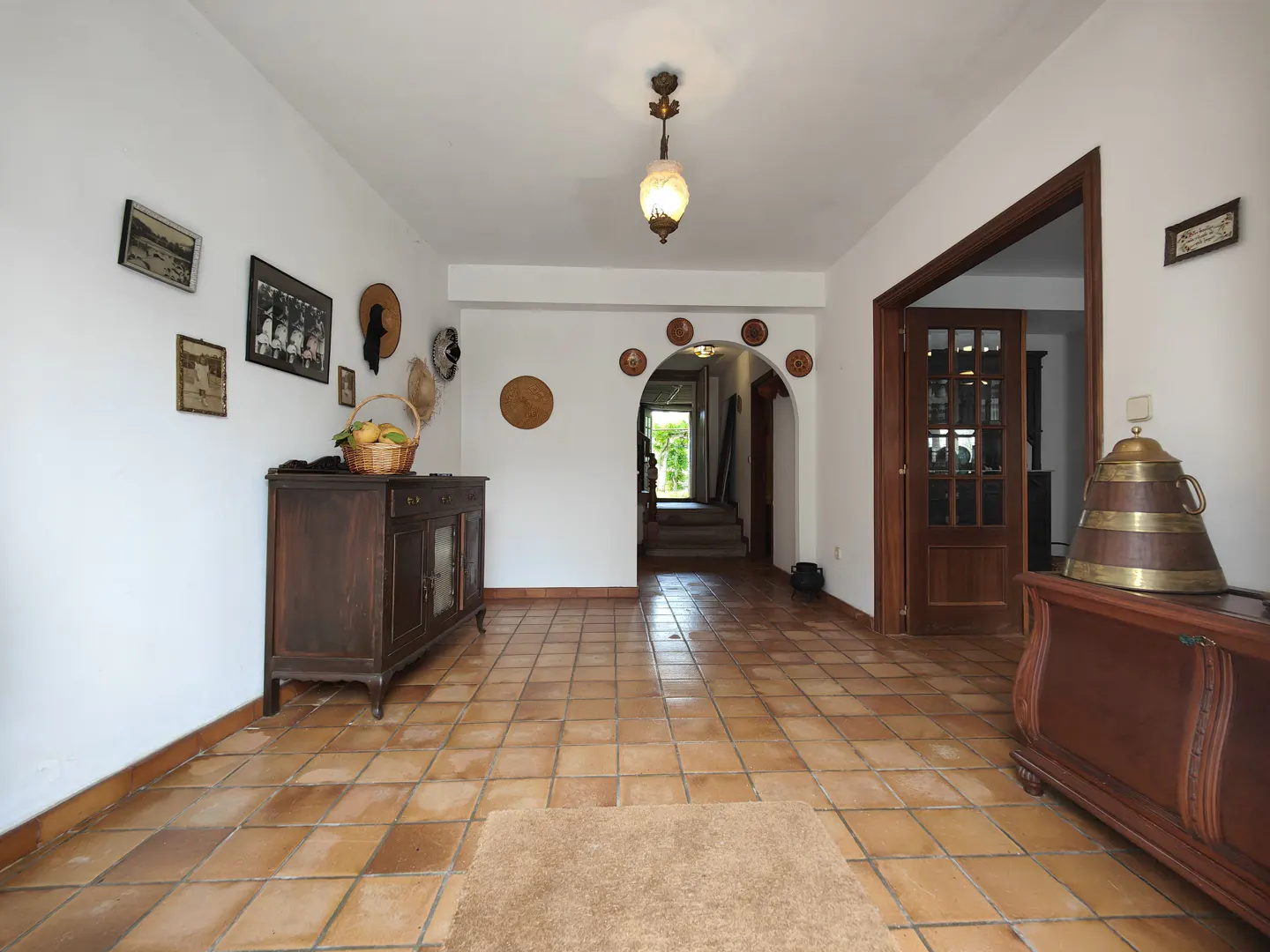 Bright foyer with terracotta tile floor, white walls, and dark wood furniture. A basket of fruit sits on a cabinet under framed photos and hats. An arched doorway leads to stairs.