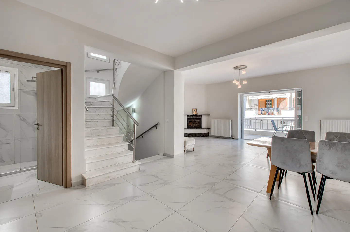 Bright, open-plan living space with marble floors, stairs, dining table with grey chairs, fireplace, and balcony access.