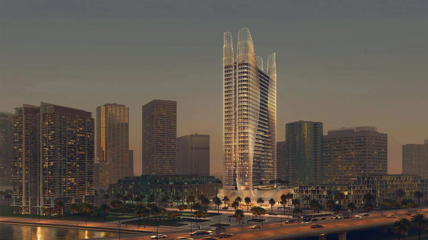 Cityscape view at dusk featuring a modern, tall skyscraper with unique architectural details, surrounded by other buildings and palm trees.