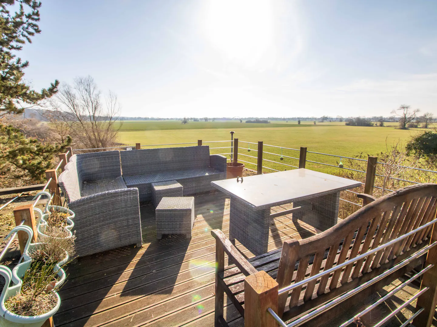 Outdoor patio with gray wicker furniture, wooden bench, and green field view. The deck has a wooden railing with metal wire. Sunny day.
