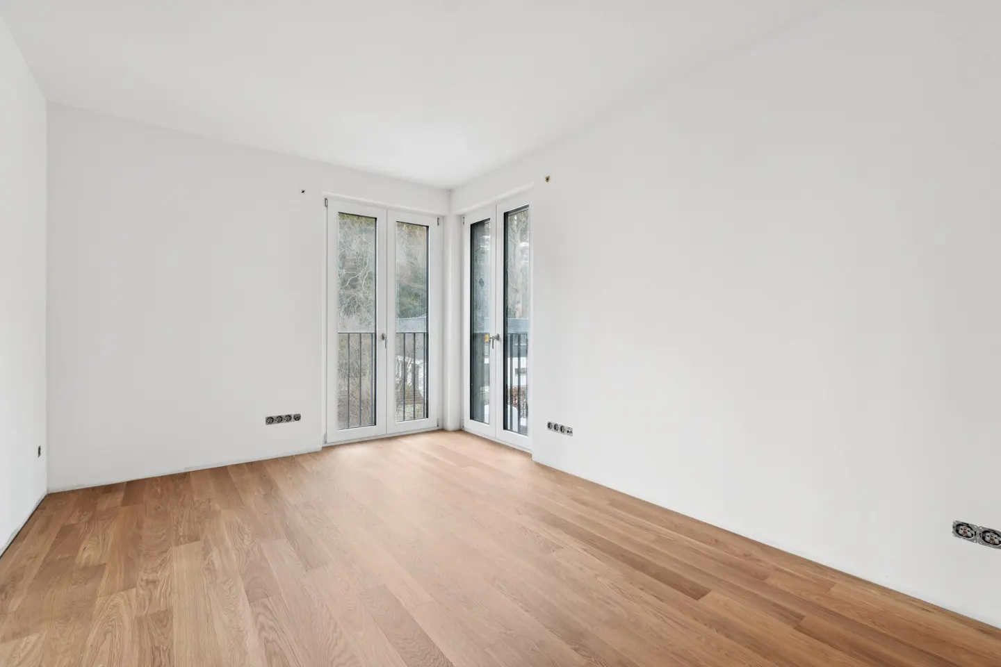 Bright, empty room with light wood floors, white walls, and double doors leading to a balcony.