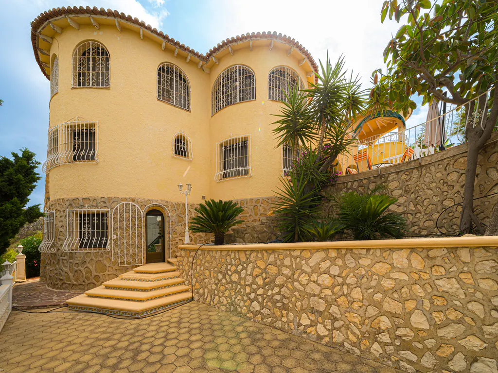 Yellow Mediterranean-style villa with arched windows, stone base, and stone-paved patio with steps leading to the entrance.