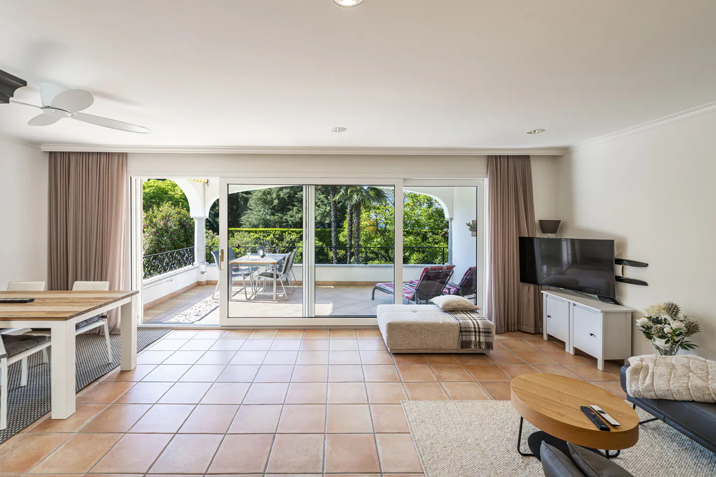 Bright living room with terracotta tile floor, dining table, TV, and sliding glass doors to a balcony with outdoor furniture.