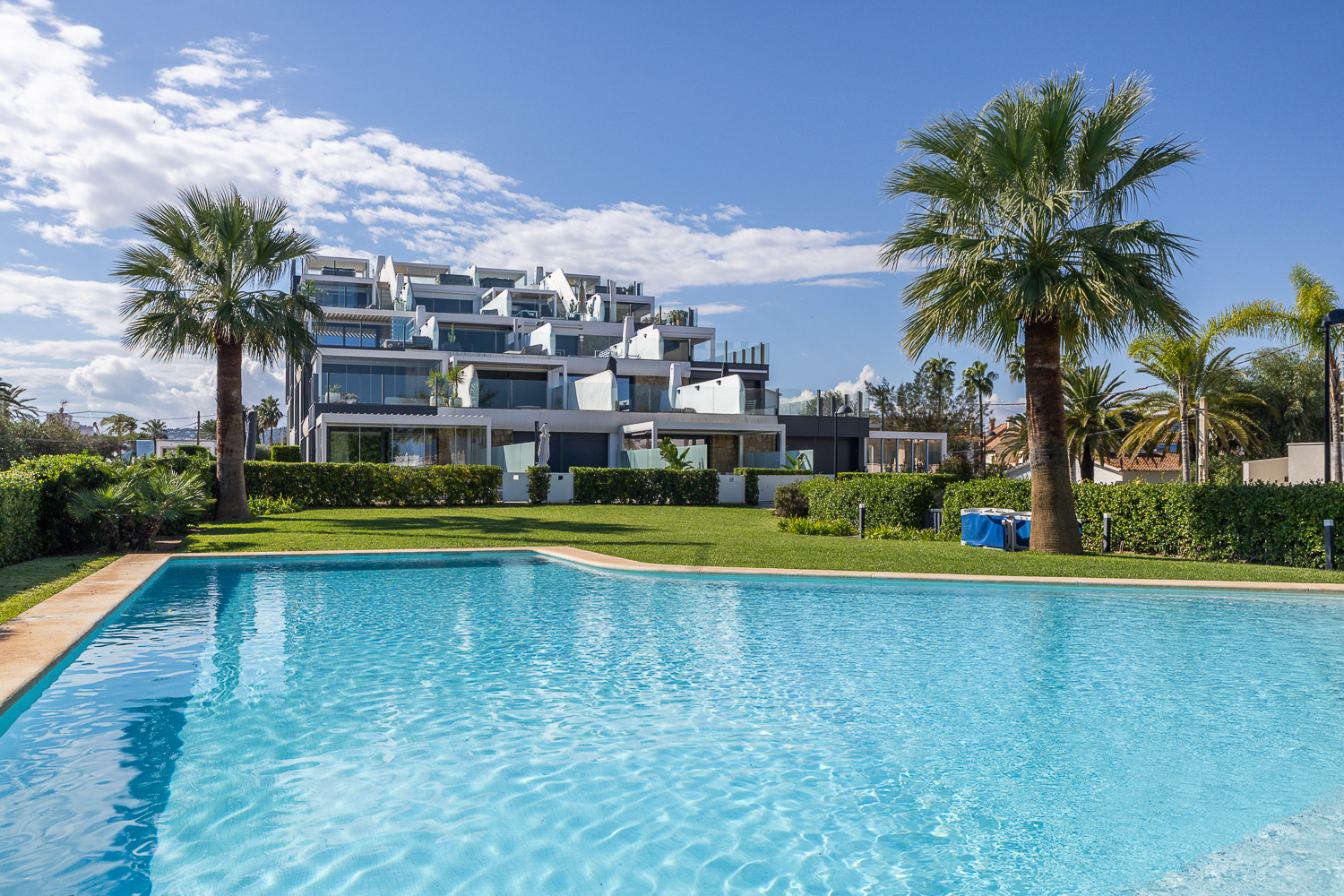 Modern white apartment building with balconies overlooking a bright blue swimming pool and green lawn under a sunny sky. [AI-generated text]