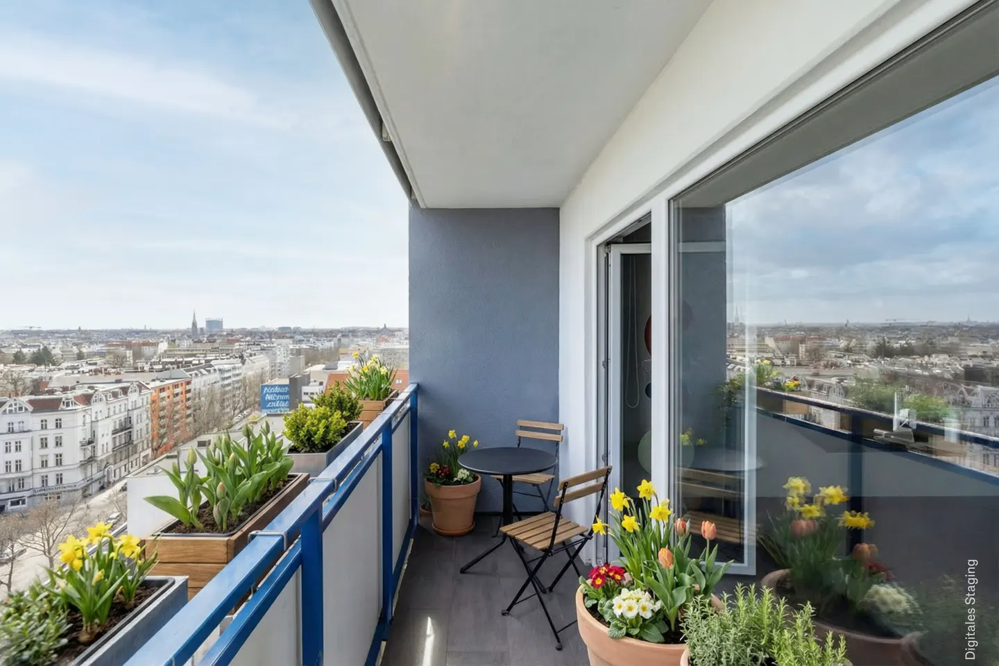 Apartment balcony with city view. Blue railing, potted flowers, and a small table with two chairs. Sliding glass doors reflect the cityscape.