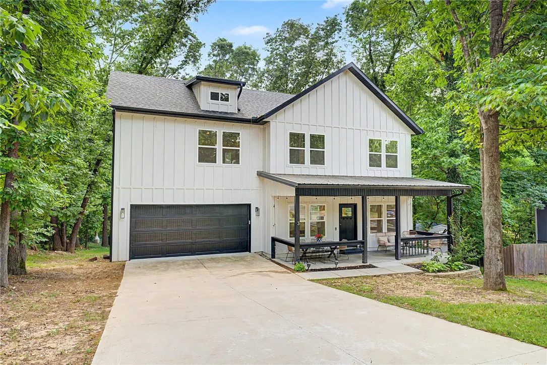 Two-story white farmhouse with black trim, a covered porch, and a dark garage door, surrounded by green trees.
