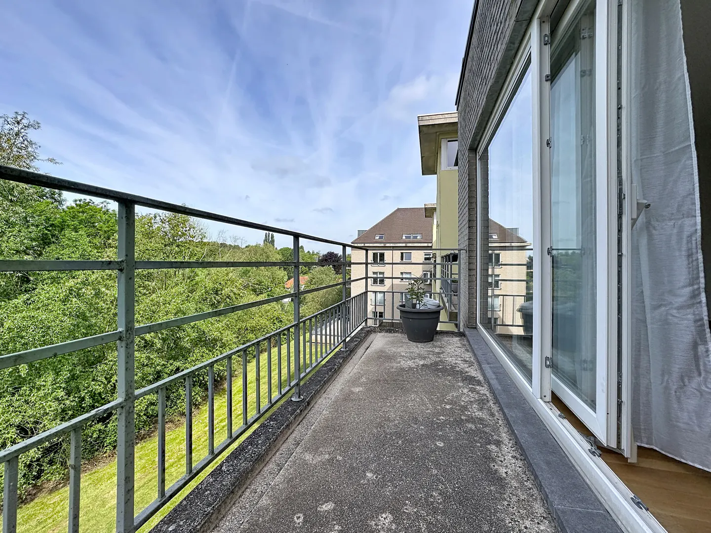 Balcony view with metal railing, concrete floor, and potted plant. Trees and a building are visible in the background under a blue sky.