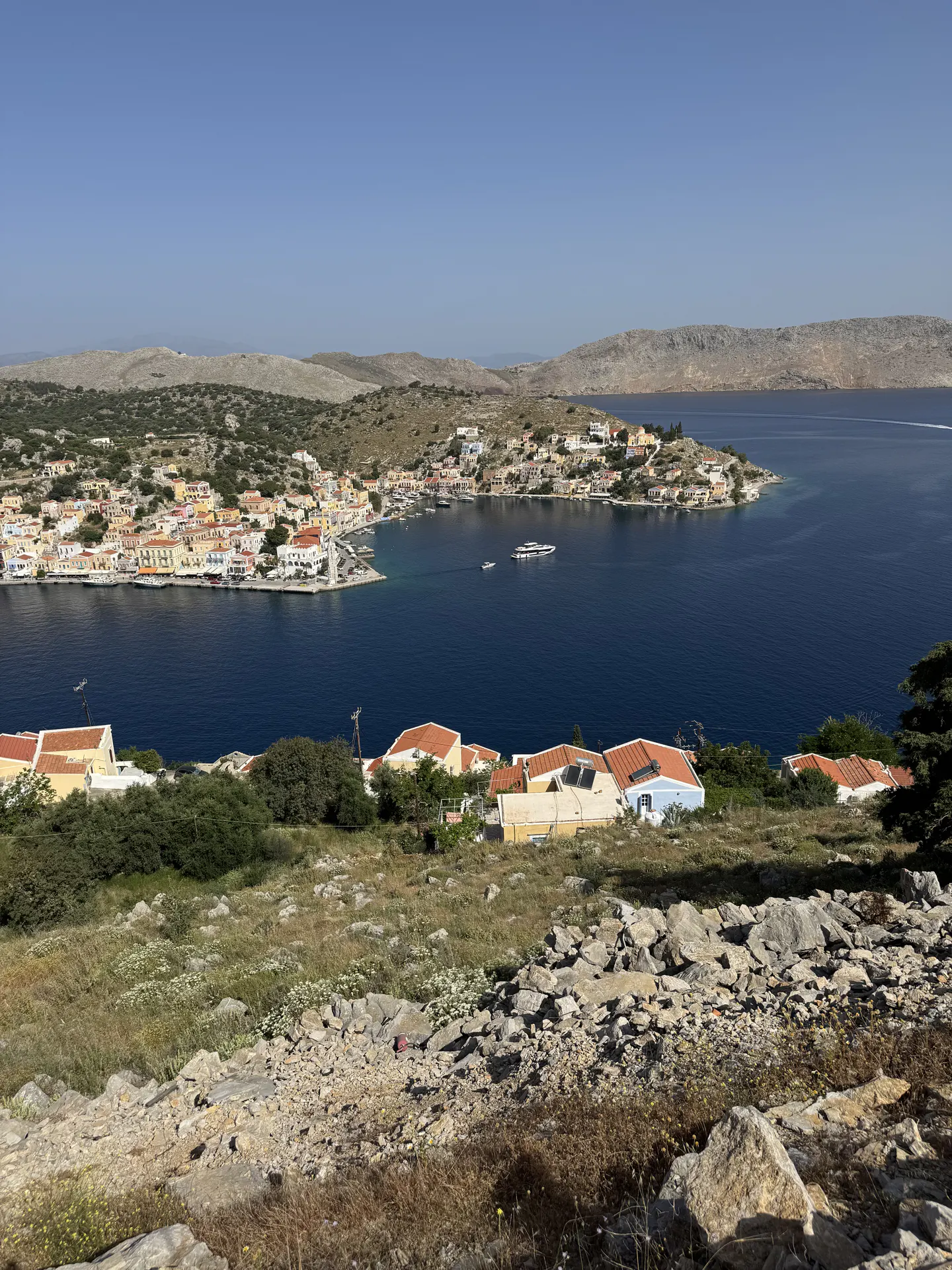 A high-angle view of a Greek coastal town with colorful houses surrounding a deep blue bay under a clear sky.