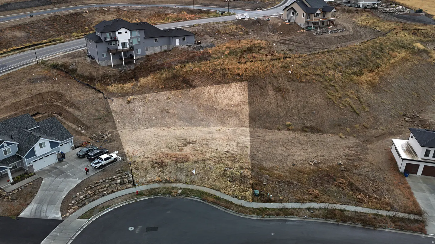 Aerial view of a vacant lot on a hill, surrounded by houses and a curved street. The lot is mostly dirt and dry grass.