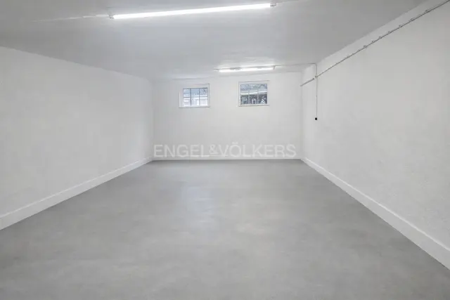 Empty, rectangular room with white walls, gray floor, and two small windows. Engel & Völkers logo on the back wall.