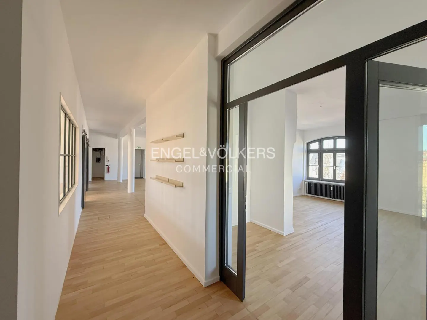 Bright office space with wood floors, white walls, and black framed glass doors. Engel & Völkers Commercial logo on the wall.
