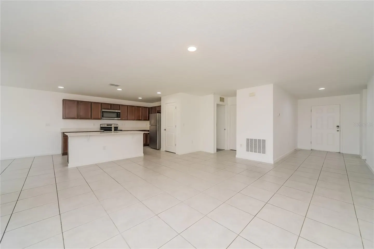 Open-concept living space with white tile floors, white walls, and a kitchen with brown cabinets and stainless steel appliances.