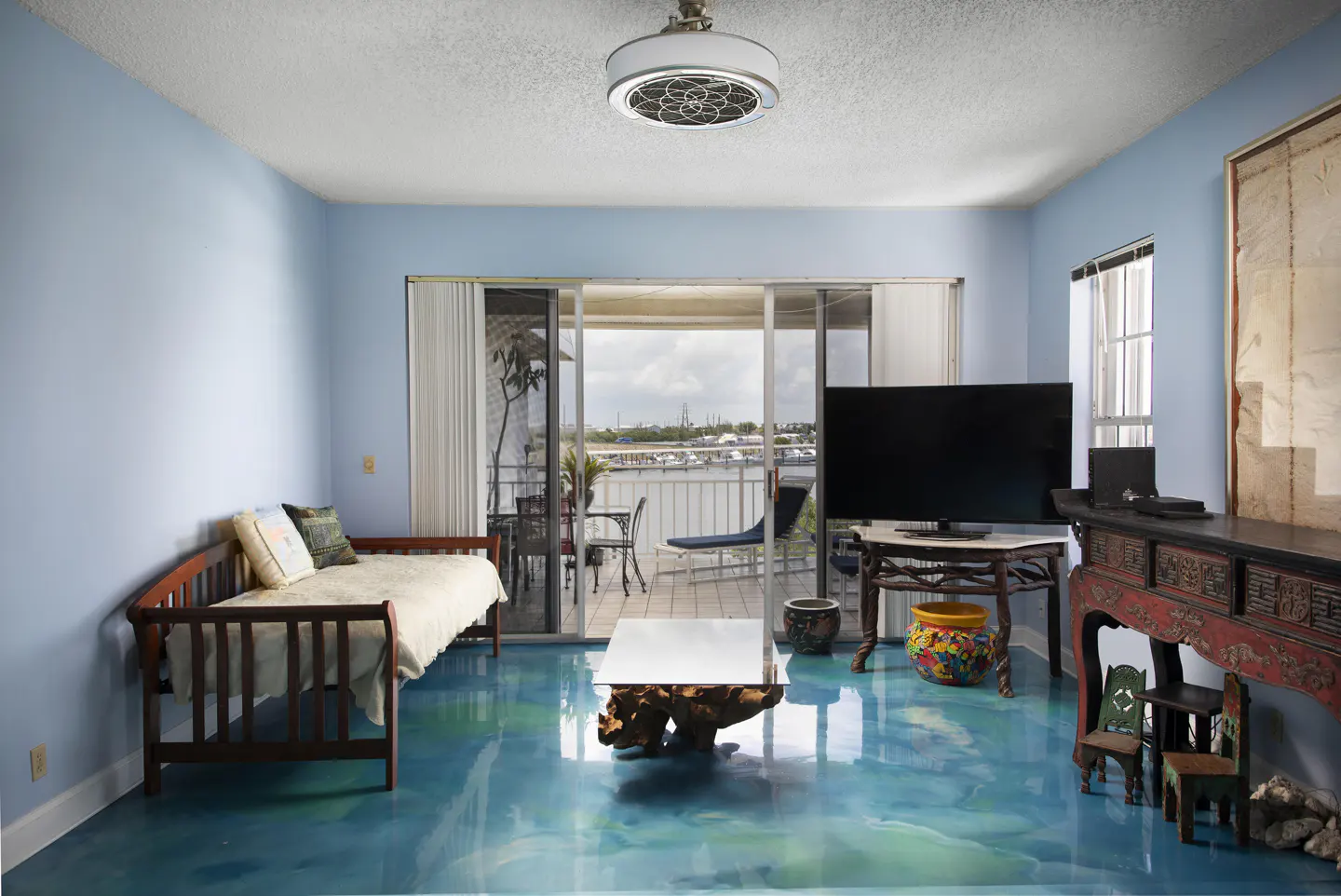 Living room with blue walls and a blue epoxy floor. A daybed, TV, and balcony with a view are visible.