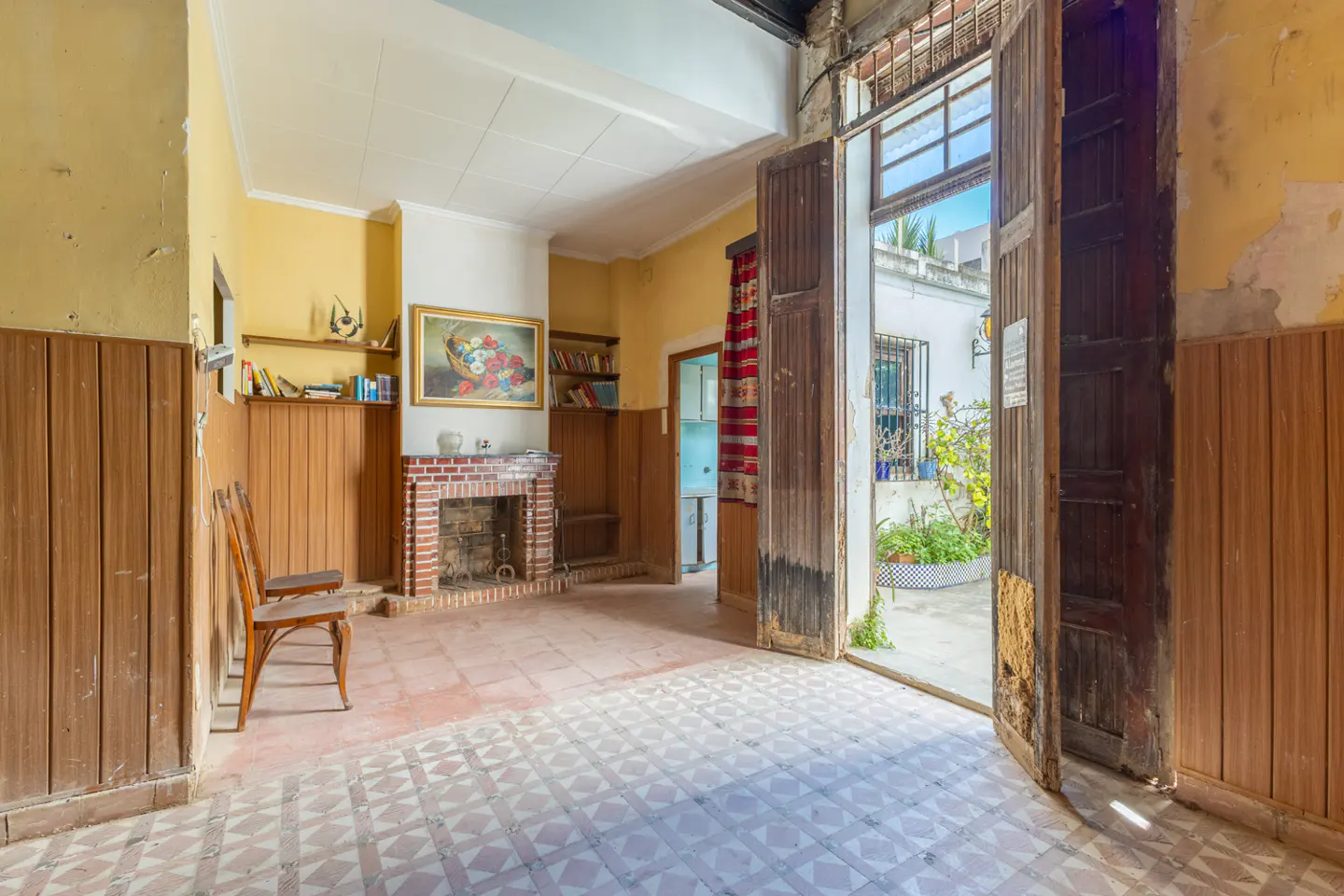 A room with yellow walls, wood paneling, a brick fireplace, and open wooden doors leading to a courtyard.