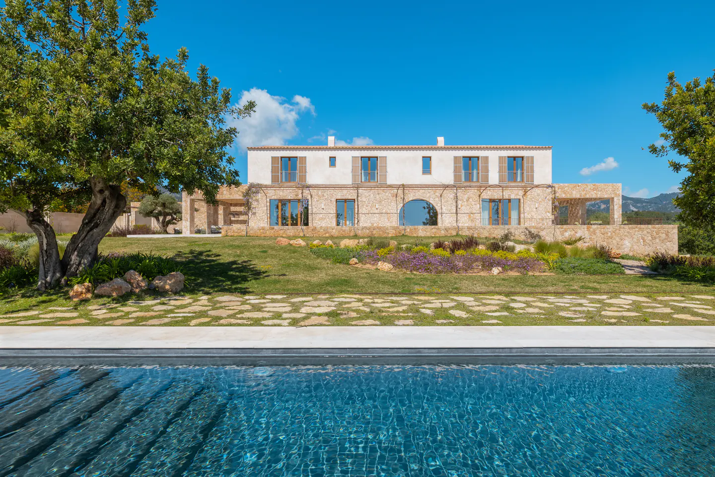 A two-story stone house with a pool in the foreground on a sunny day. The house has white walls on the second story and brown shutters.