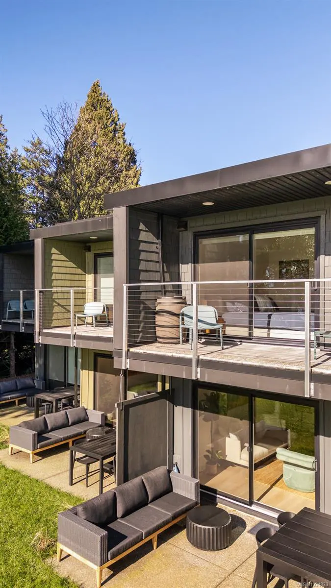 Modern two-story building with outdoor seating on patios and balconies. Gray sofas and tables are on the ground level. Balconies have chairs and barrels.