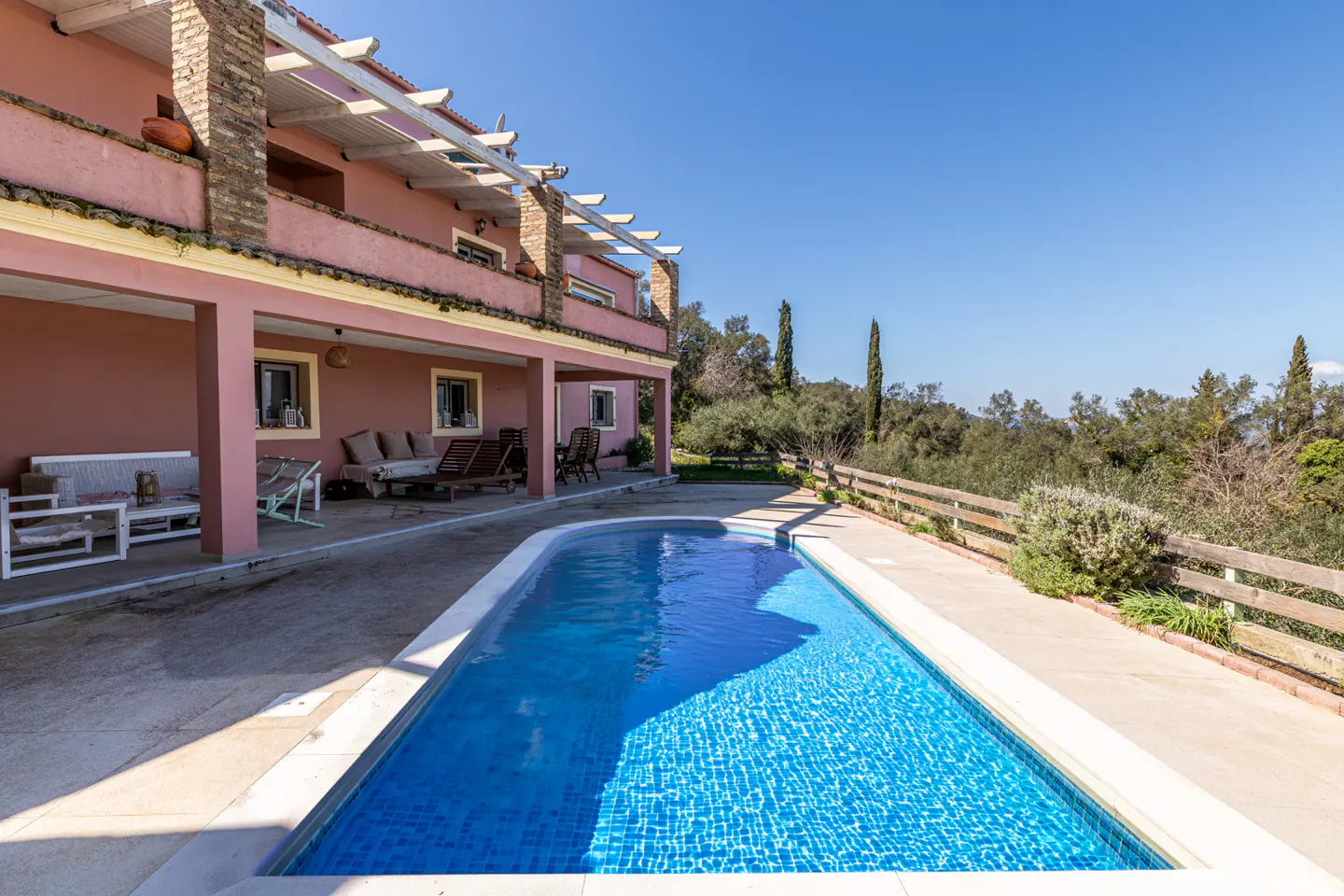 A pink two-story house with a blue tiled pool in the foreground on a sunny day.