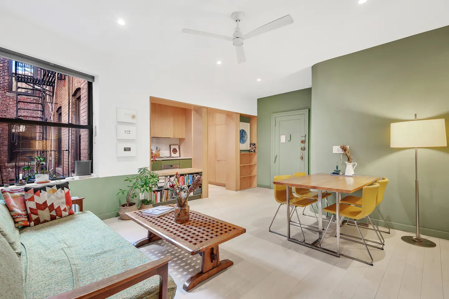 A bright living room with a green sofa, wooden table, and yellow chairs. A window shows a brick building with a fire escape.