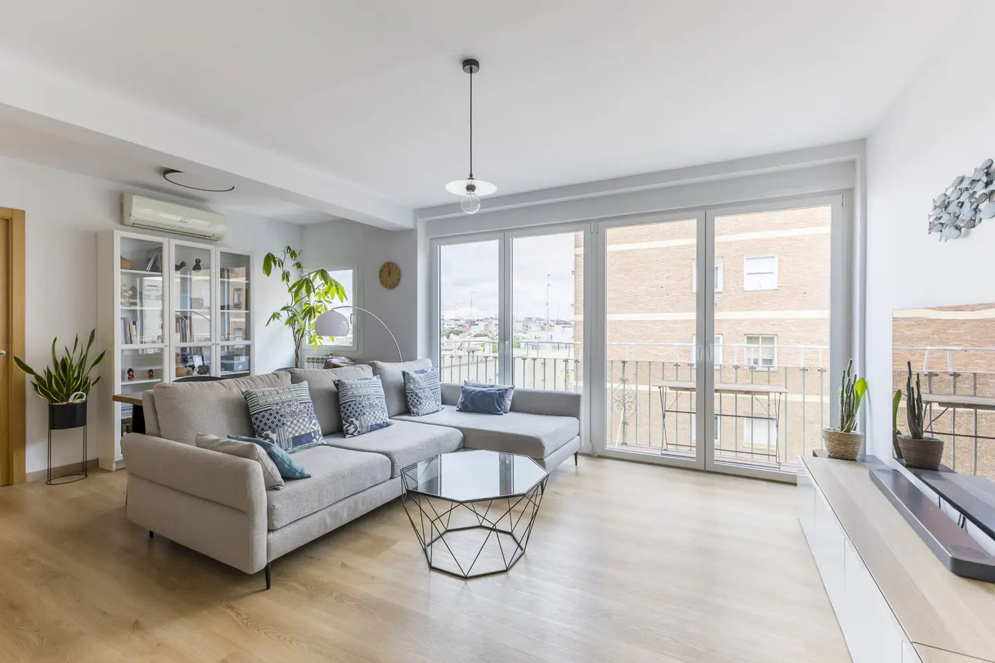 Bright living room with a gray sectional sofa, patterned pillows, and a glass coffee table. Balcony doors offer city views.