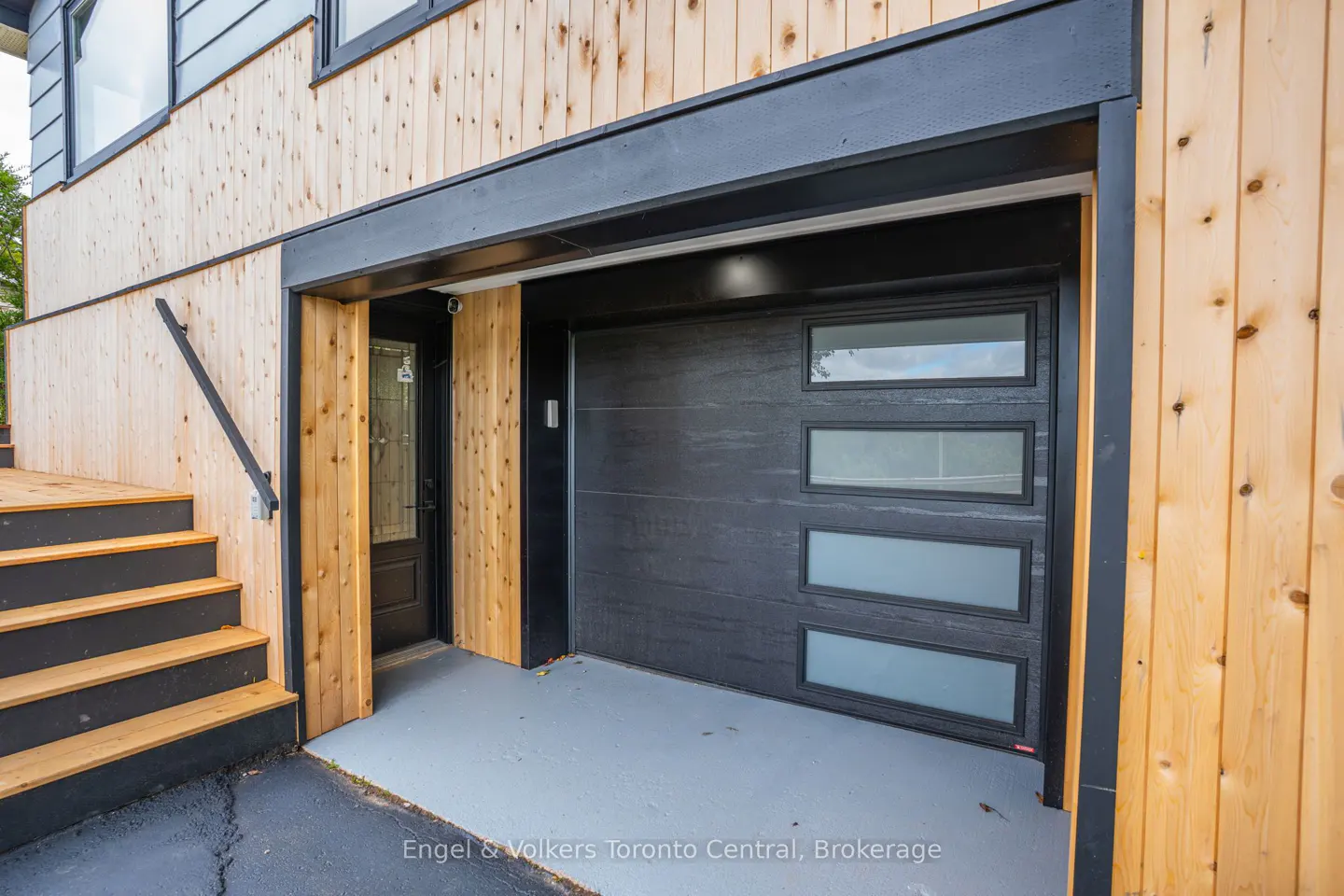 Exterior view of a modern home with light wood siding, a black garage door with windows, and black-trimmed stairs.