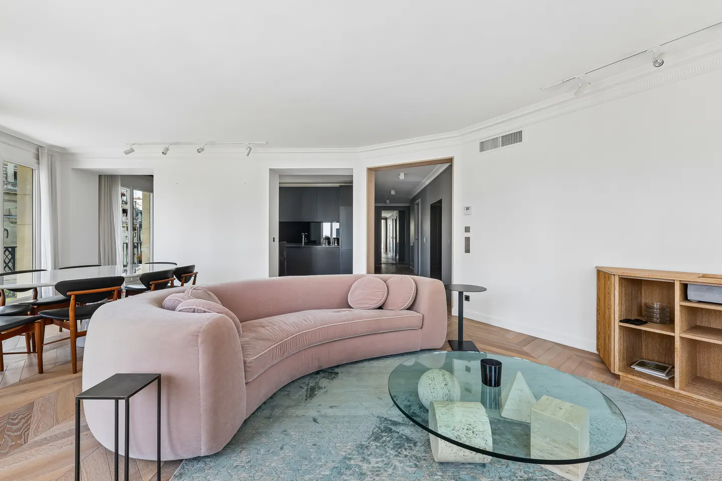 Modern living room with a curved pink velvet sofa, glass coffee table, blue area rug, and wooden shelving on parquet floors.