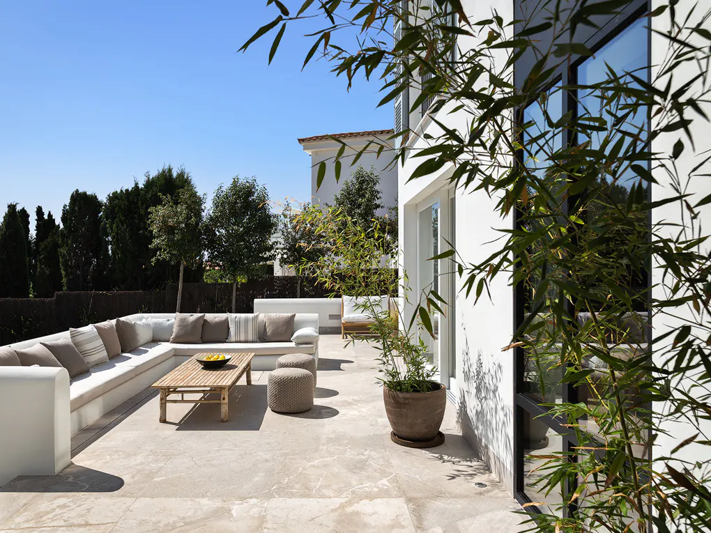 Outdoor patio with white sectional sofa, wood table, and potted plant. Blue sky and green trees in the background.