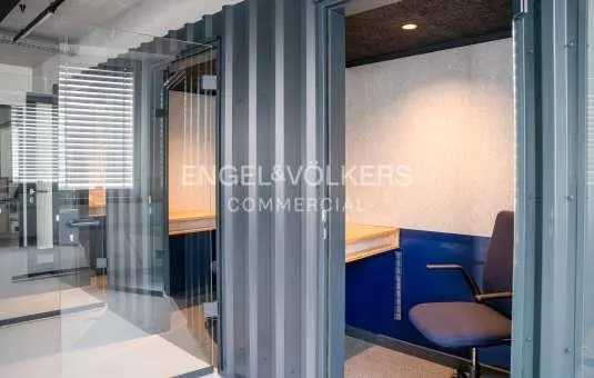 Office space with a blue chair and desk inside a gray corrugated metal booth, seen through a glass wall.