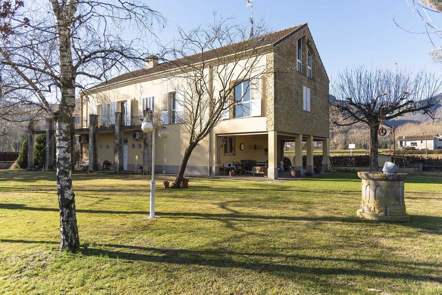 Two-story yellow house with a large lawn, trees, and a stone well in the foreground on a sunny day.