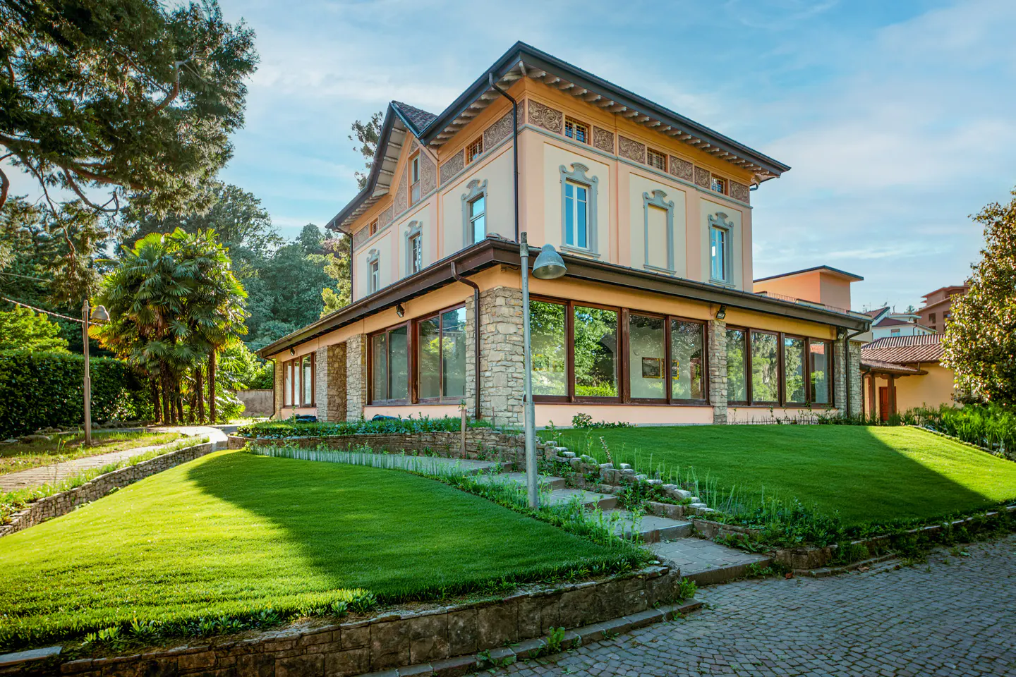 Two-story peach-colored villa with stone base, large windows, and a manicured lawn under a bright blue sky.