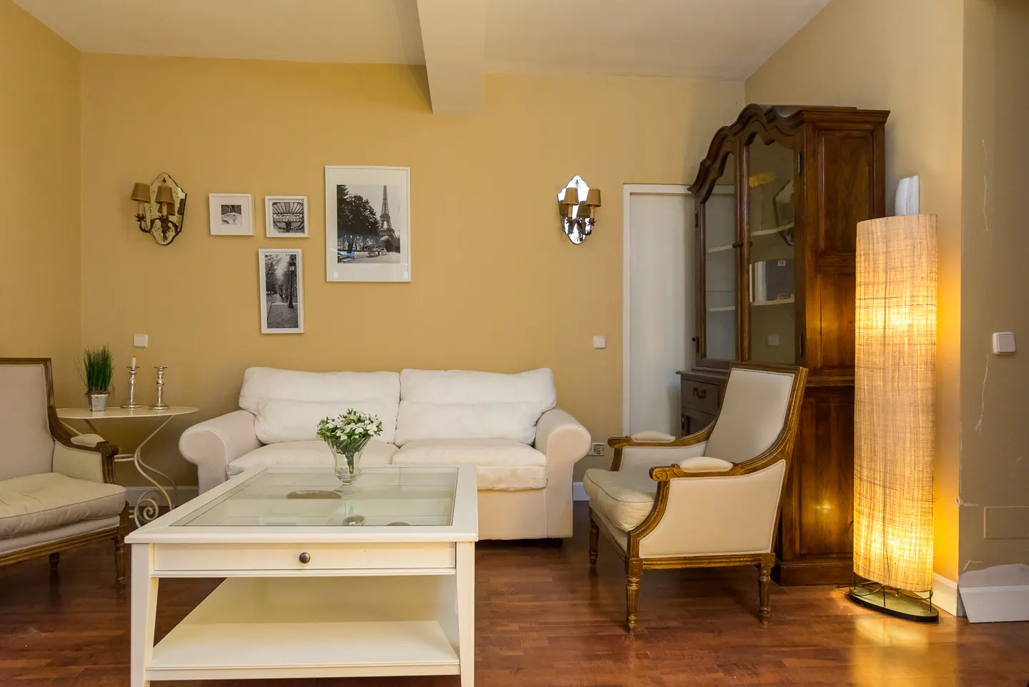 Living room with white sofa, glass coffee table, and antique cabinet on wood floor. Walls are yellow with framed art.