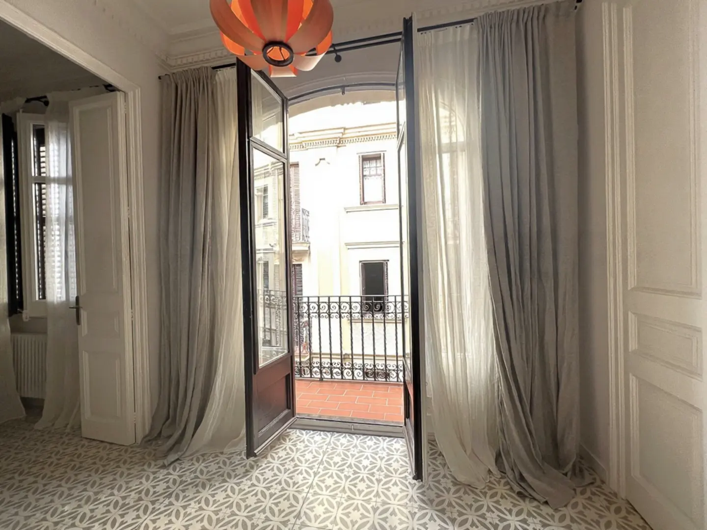 Bright room with patterned tile floor, open French doors to balcony. Gray and white curtains frame the doors. Orange pendant light.