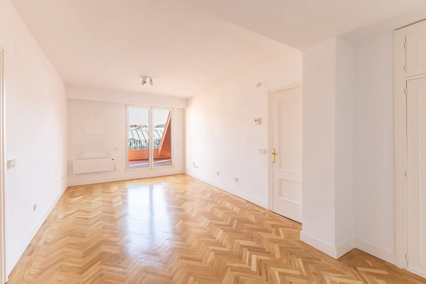Bright, empty room with herringbone wood floors, white walls, and a window to a balcony.