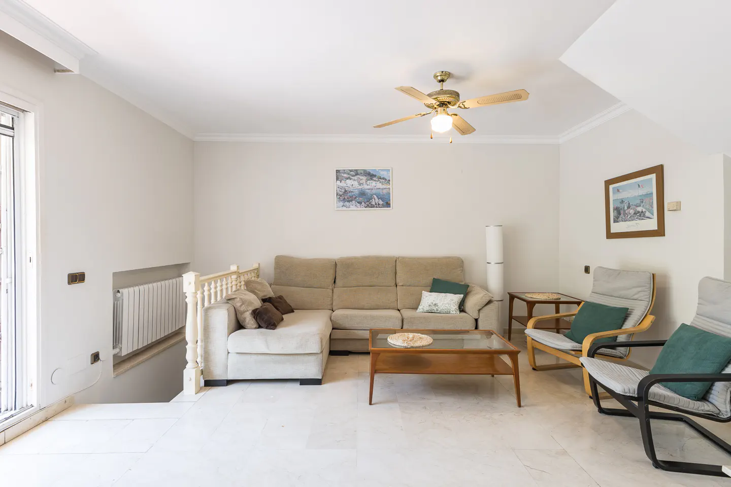 Bright living room with beige sectional sofa, two armchairs, and a glass-topped coffee table. A ceiling fan hangs above.