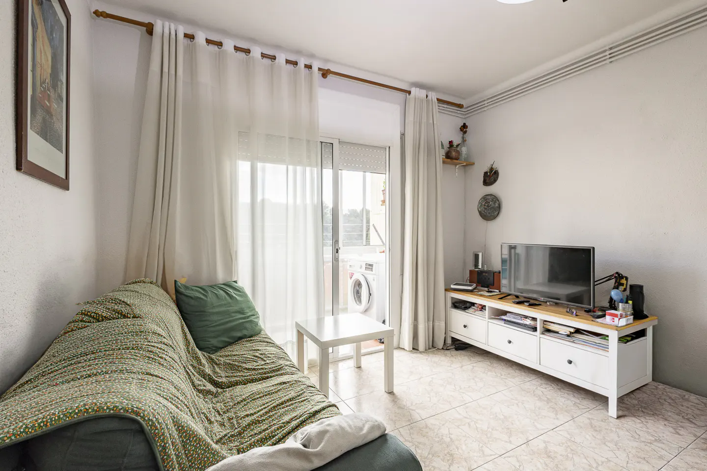Living room with a green sofa, white curtains, and a TV stand. A white table sits in front of the sliding glass doors to a balcony.