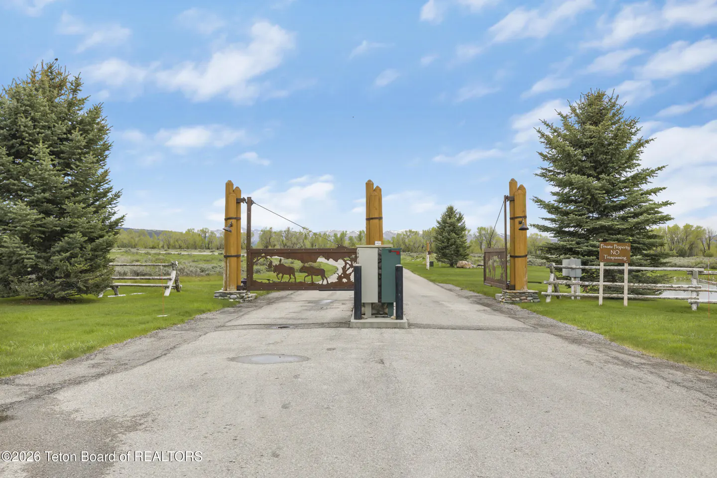 Gated driveway entrance to a property. The gate has a metal moose design and wooden posts. Green grass and trees surround the driveway.