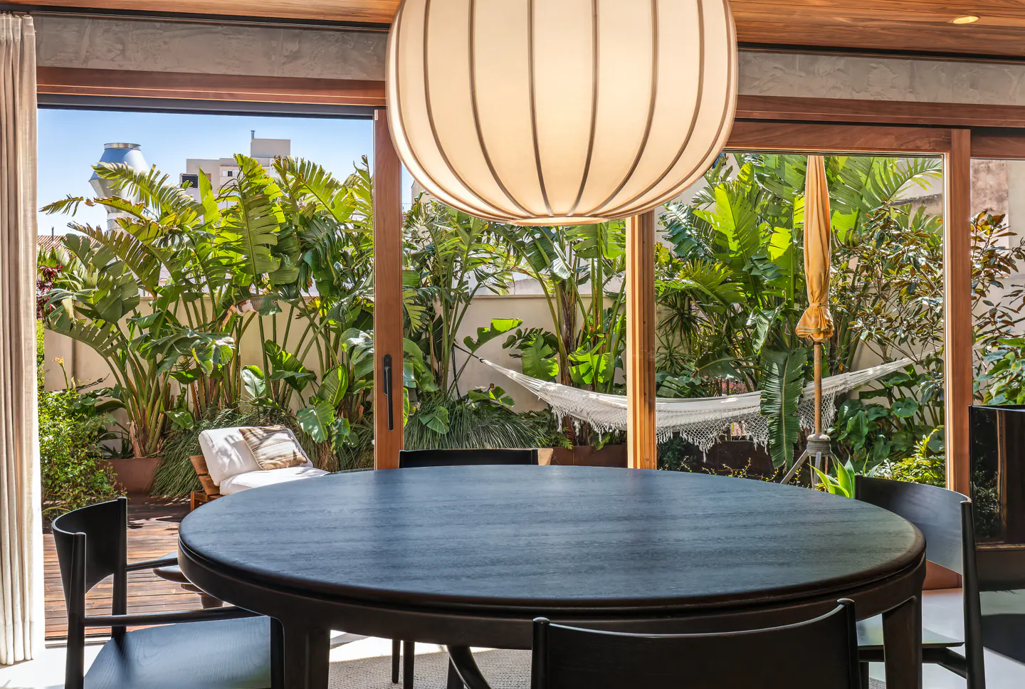 Indoor dining area with a round table and black chairs, overlooking a lush outdoor patio with a hammock and lounge chair.