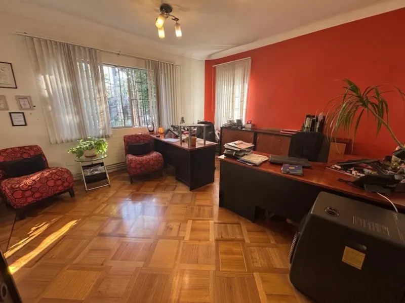 Office space with wood floors, red accent wall, and two desks. Two red patterned chairs sit near a window with white curtains.