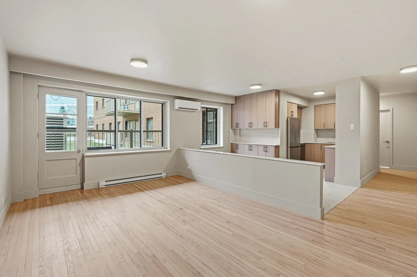 Bright, open apartment with hardwood floors, a kitchen with light wood cabinets, and large windows overlooking a brick building.