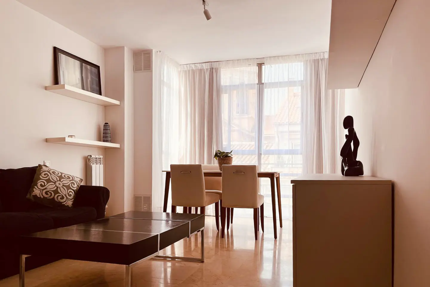 Living room with a black sofa, a dark wood coffee table, and a dining table with four chairs. White walls and curtains.