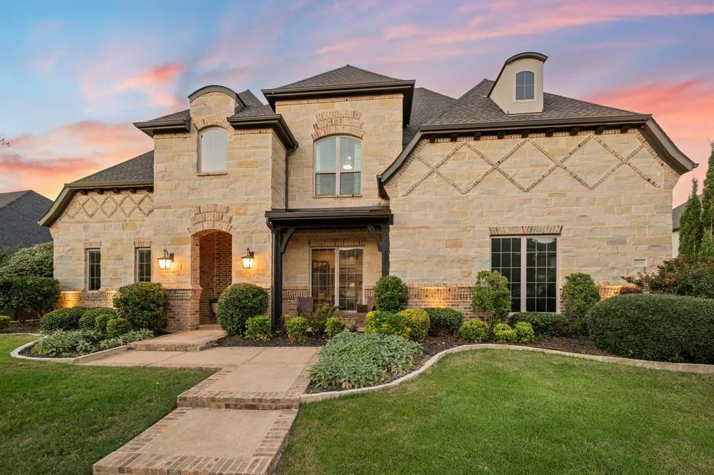 A two-story stone house with a dark roof, green lawn, and landscaped bushes under a colorful sunset sky.