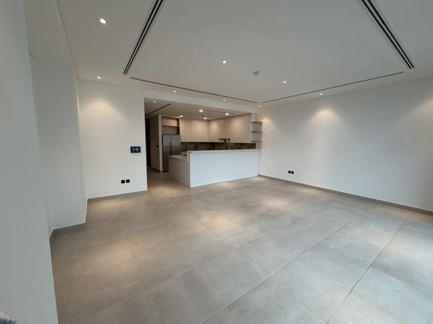 An open-concept living space with white walls, gray tile floors, and a kitchen in the background. The kitchen has white cabinets and a breakfast bar.