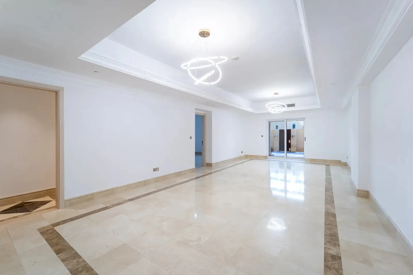 Bright, empty room with white walls, marble floors, and modern light fixtures. Sliding glass doors lead to an outdoor area.