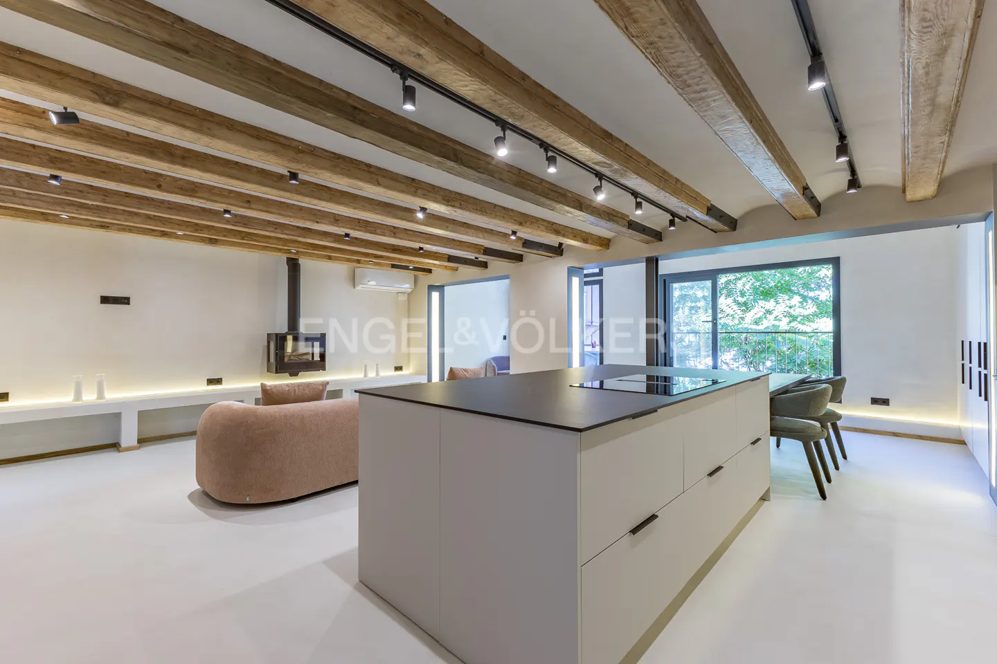 A modern, open-concept living space with exposed wooden beams, track lighting, and a large kitchen island. A fireplace and sofa are visible in the background.