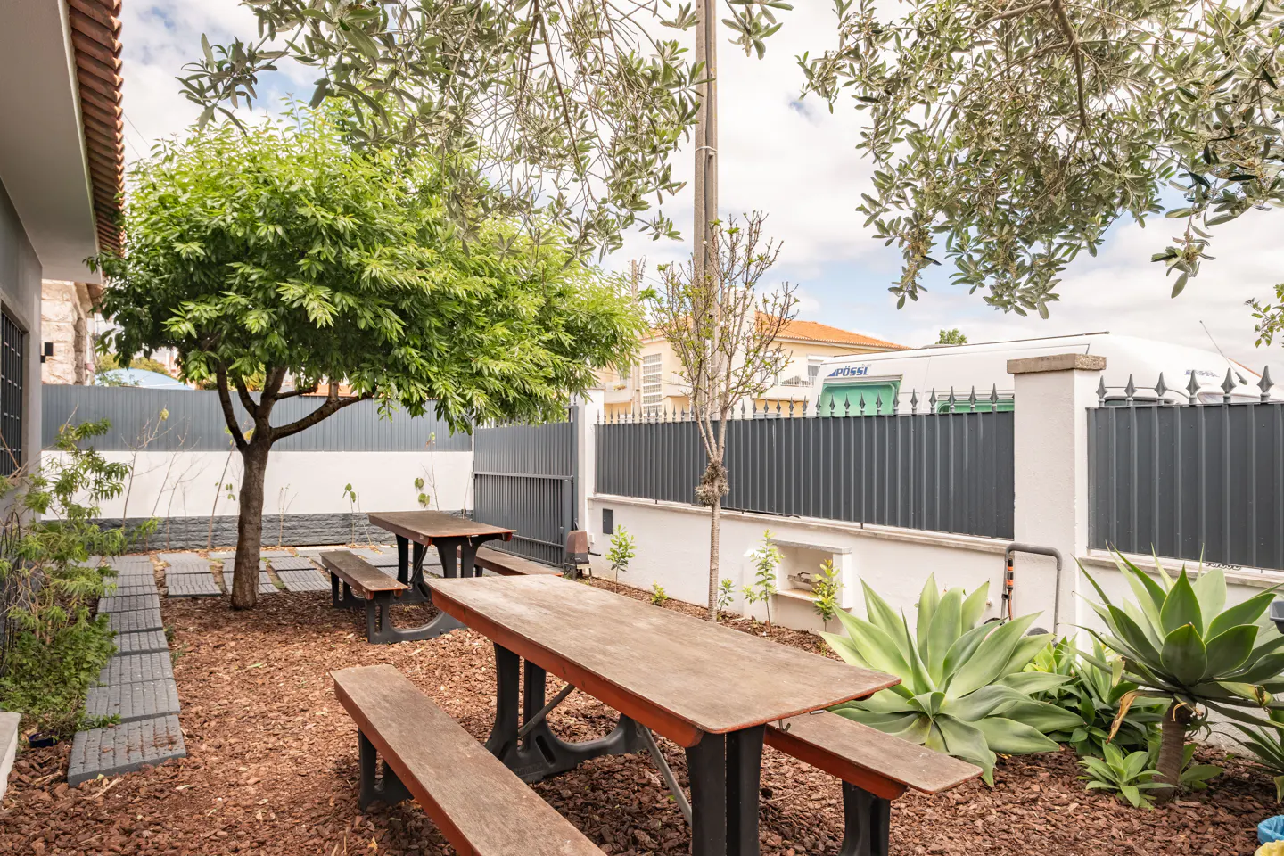 Outdoor seating area with wooden picnic tables and benches on a mulch ground, surrounded by trees, plants, and a gray fence.