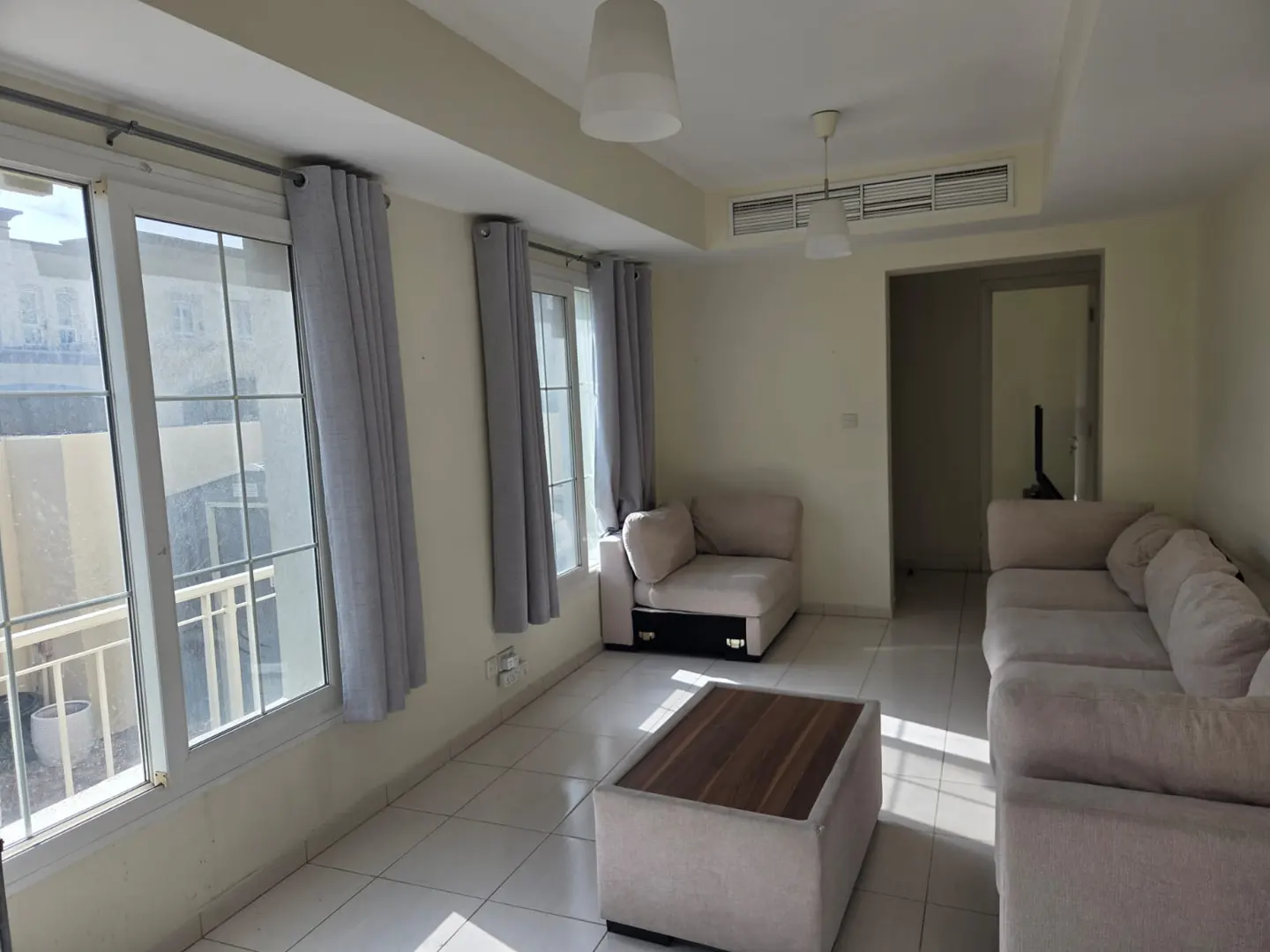 A bright living room with white tile floors, beige walls, and large windows with gray curtains. A beige sectional sofa and a coffee table are in the center.