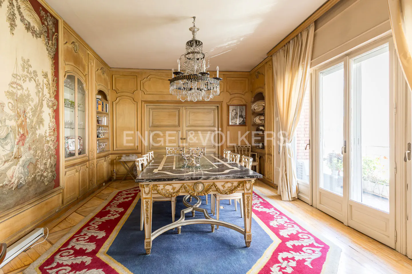 Ornate dining room with a marble-topped table, crystal chandelier, and tapestry wall art. Light wood paneling and a blue rug with red trim add warmth.