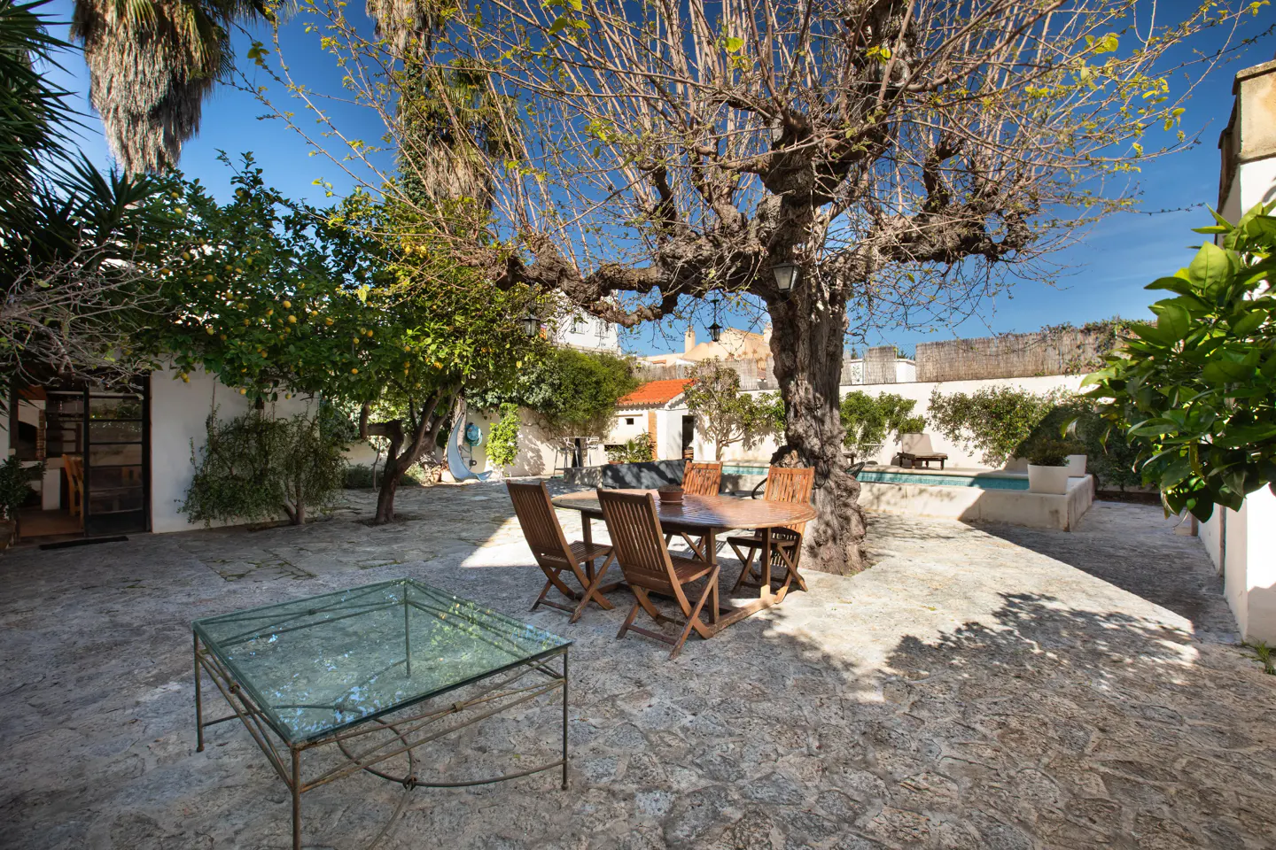 Outdoor patio with stone flooring, table and chairs under a large tree. Swimming pool and white walls in the background.