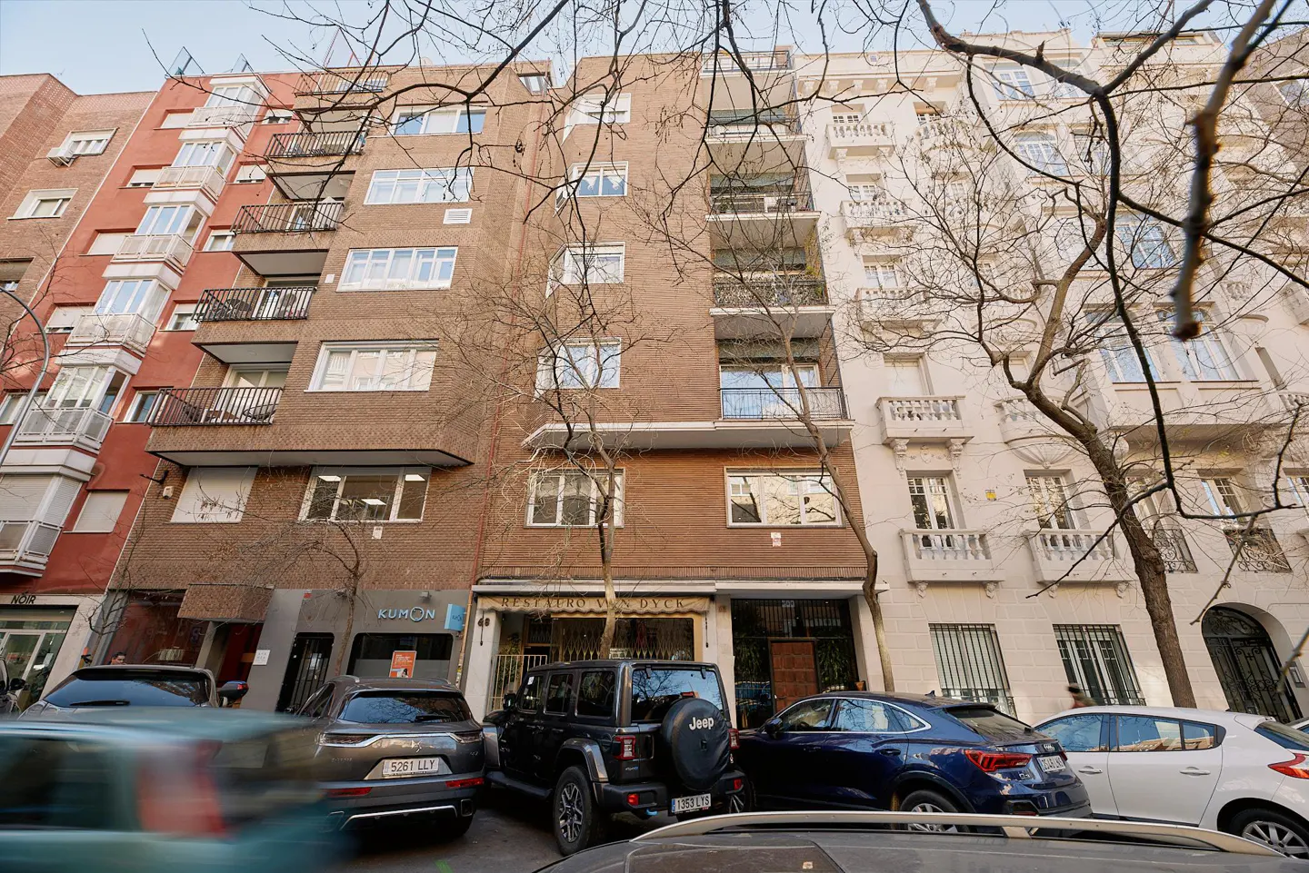 Street view of a multi-story apartment building with balconies. Cars are parked in front of the building. Bare trees are in the foreground.