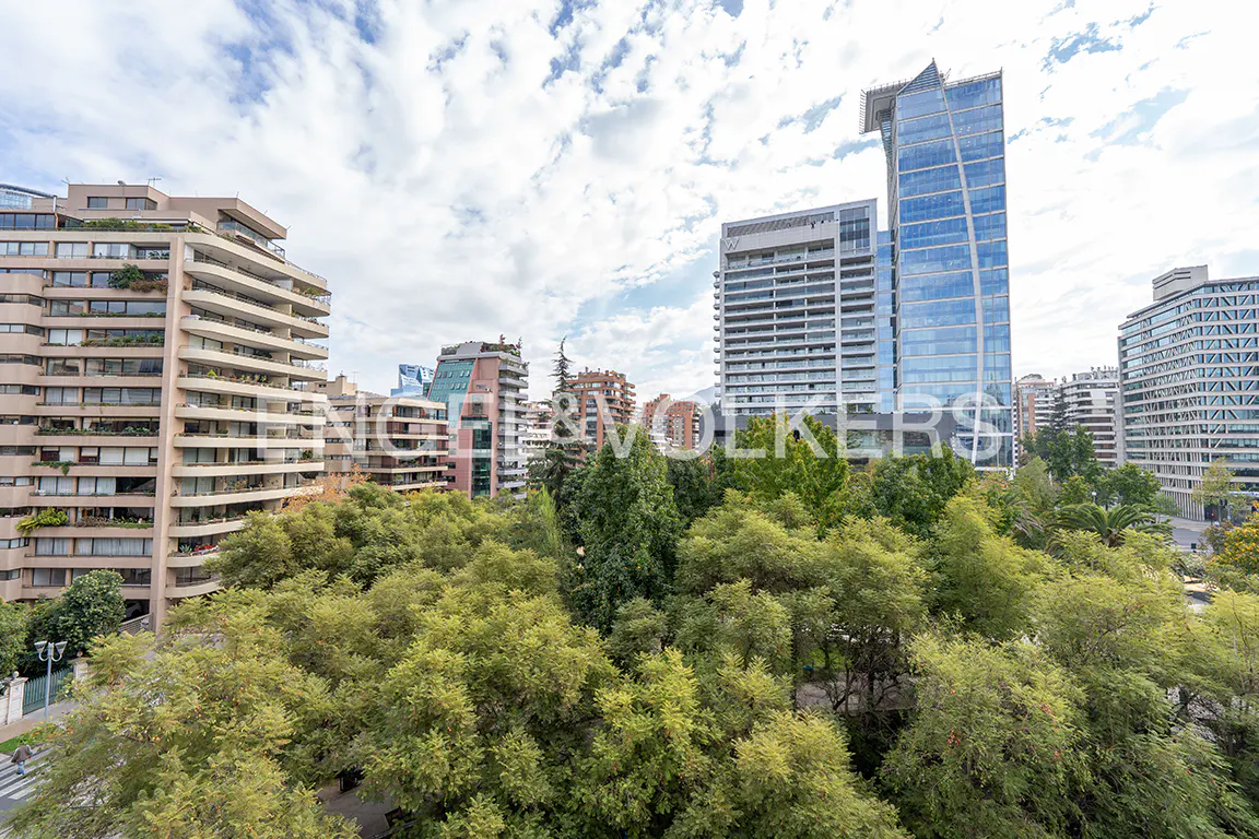 Cityscape view with green trees in the foreground and tall buildings in the background under a cloudy sky.